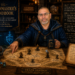 A cinematic Dungeons and Dragons inspired scene featuring Frank Jamison portrayed as a wise guildmaster mentor seated at a wooden strategy table inside a candlelit guild hall. Wearing dark blue robes, Frank studies a fantasy campaign map symbolizing a software developer career journey, with miniature adventurers marking paths through Choosing Your Path, Surviving the Guild Hall, The Trials of the Realm, and Becoming the Mentor. Glowing code appears on a magical screen nearby alongside dice, books, candles, and developer themed artifacts. A large banner displays The Guildmaster’s Handbook: Becoming the Developer Everyone Wants on the Quest with the tagline Code wins battles. Wisdom wins campaigns. The atmosphere feels warm, wise, and adventurous, blending software development with D&D mentorship.